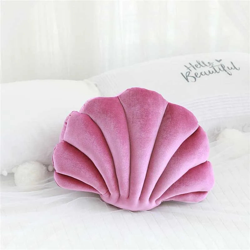 A pink velvet stuffed sofa cushion shaped like a decorative seashell resting on white bedding.