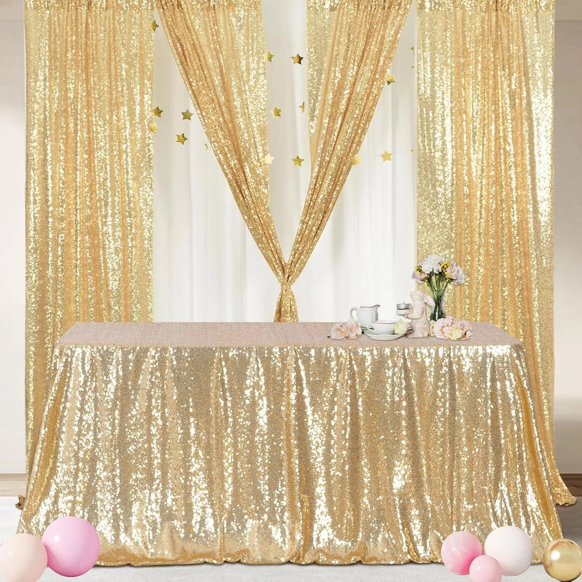A gold sequin rectangle table cover draped with sheer gold curtains and decorated with pink flowers.