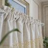 A close-up of a rustic crochet half curtain with linen weave in cream and beige tones hanging indoors.