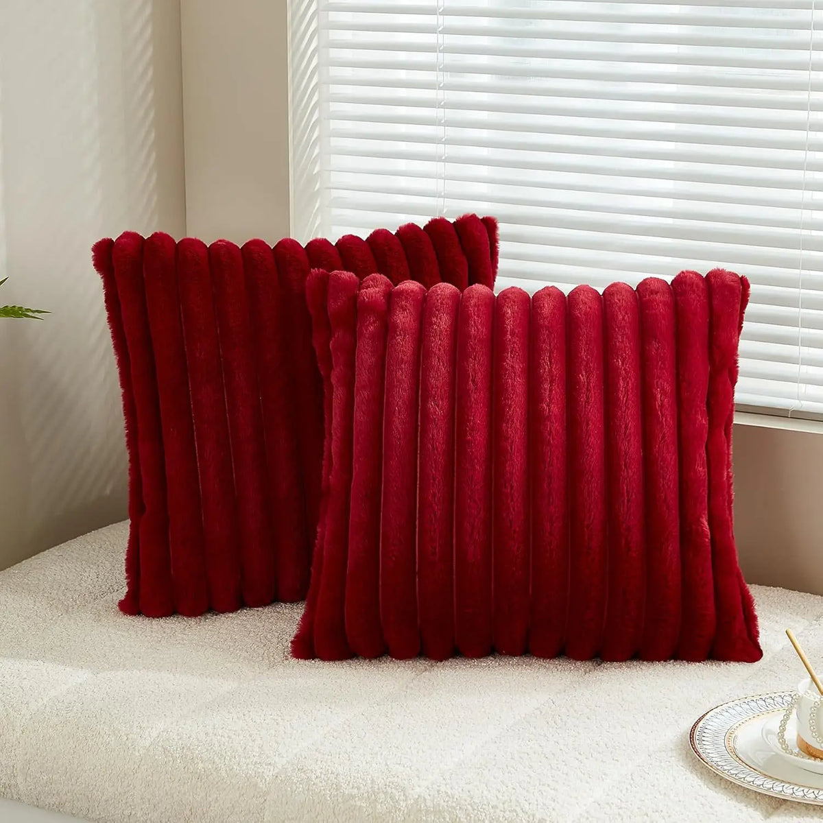 A pair of deep red faux rabbit fur cushion covers resting on a light-colored surface next to a white window blind.