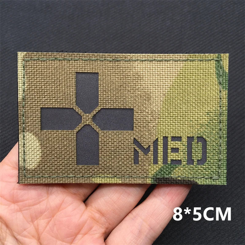 A rectangular tactical patch with a dark cross symbol on a camouflage background is held in a hand.