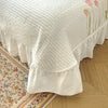 White quilted bed skirt with pink and green floral embroidery on a wooden floor.