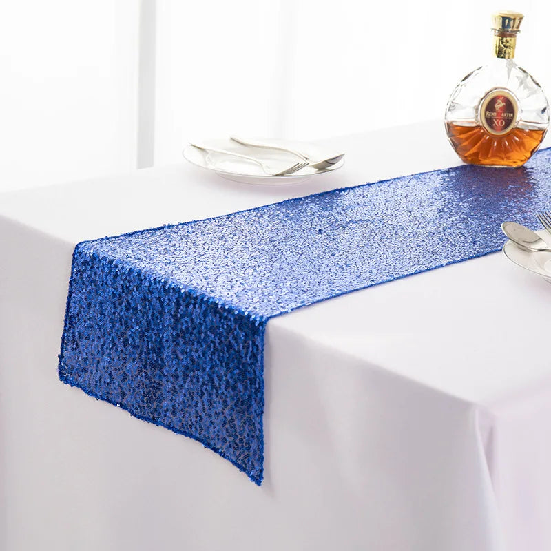A blue sequin table runner is displayed on a white tablecloth next to a bottle of amber liquid.
