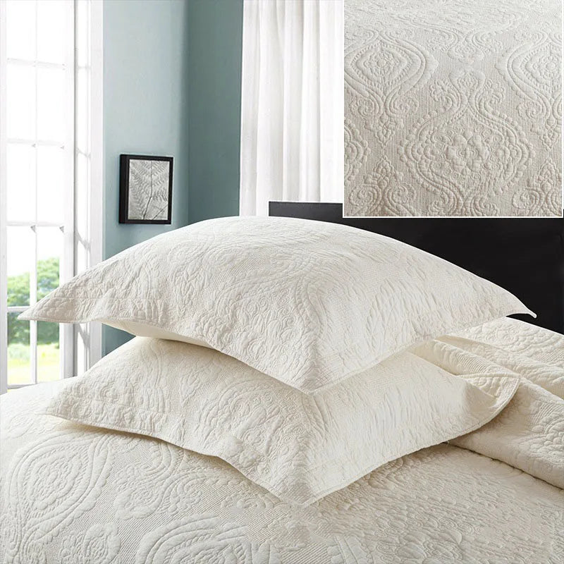 A set of white embroidered cotton quilted pillow covers displayed on a bed with sheer curtains and a framed artwork.