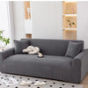 A gray stretchable full-wrap sofa slipcover with a light-colored decorative pillow and magazines on the floor.