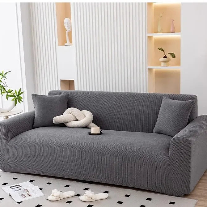A gray stretchable full-wrap sofa slipcover with a light-colored decorative pillow and magazines on the floor.