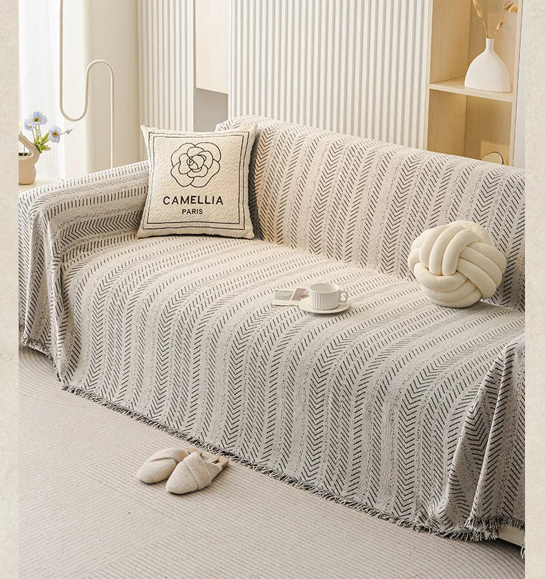 A beige sofa cover featuring an elegant fishbone pattern, decorated with a cream pillow and a white knitted sphere.