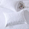 A white embroidered cotton quilted pillow cover set resting on a white textured surface with a small glass and wooden stick.