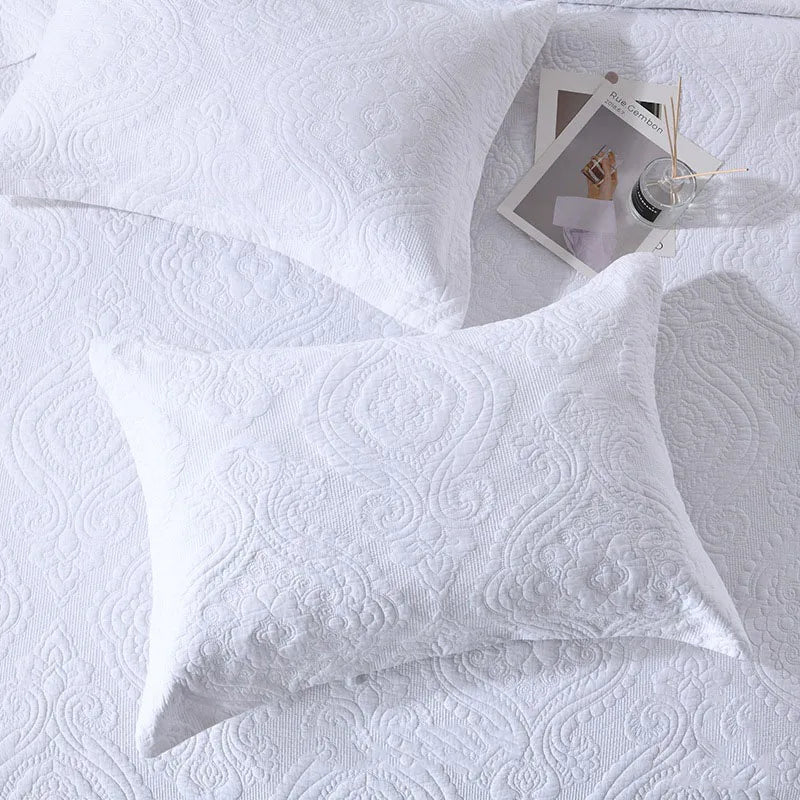 A white embroidered cotton quilted pillow cover set resting on a white textured surface with a small glass and wooden stick.