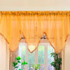 A sheer yellow curtain with heart lace and tassels hangs over a white window frame.