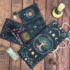 A dark fabric pouch decorated with a mystical tree of life design and crescent moons, resting on a wooden surface.