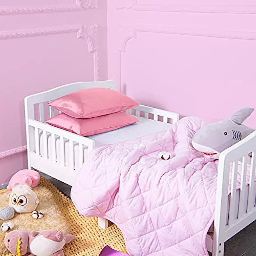 A pink quilted blanket and pillows are displayed on a white wooden crib with a pink wall background.