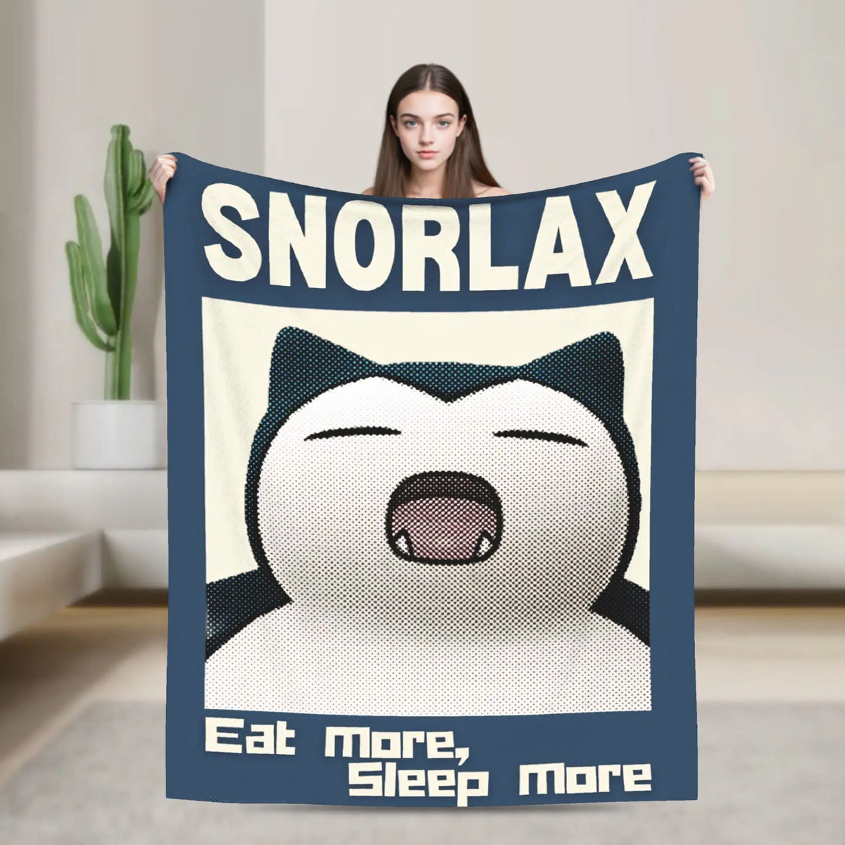 A person holds up a dark blue blanket featuring a white cartoon face and the text SNORLAX.