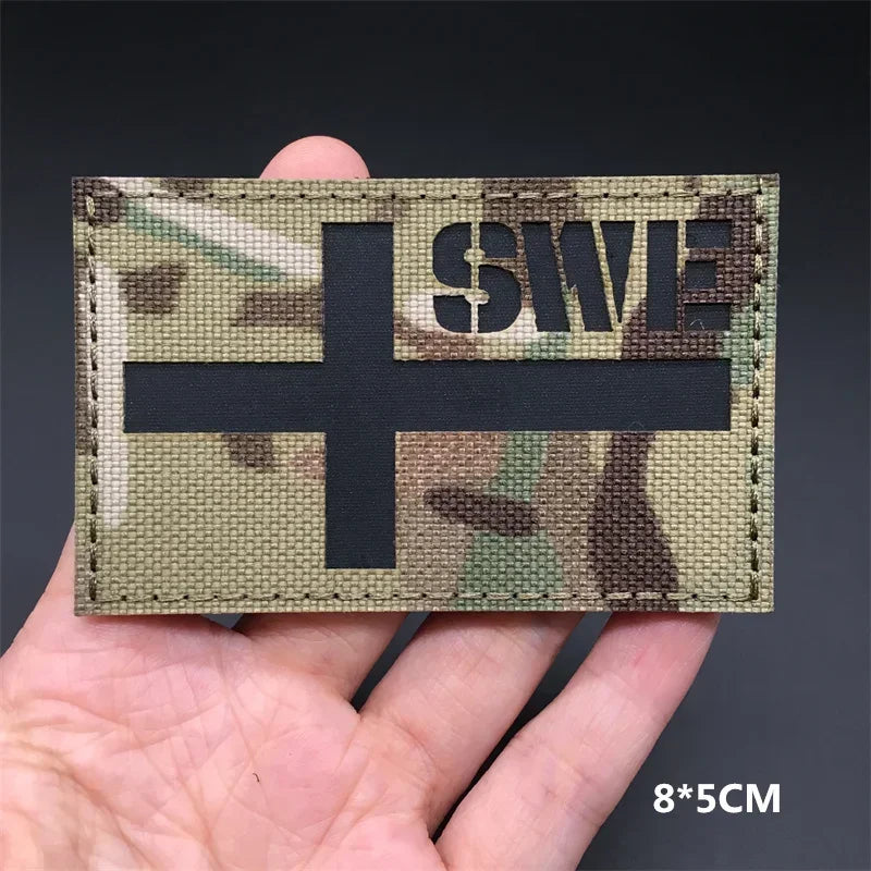 A rectangular tactical patch featuring a camouflage pattern with a black cross and the text SWE.