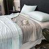 A light blue reversible cooling quilt is displayed on a bed with gray bedding and pillows.