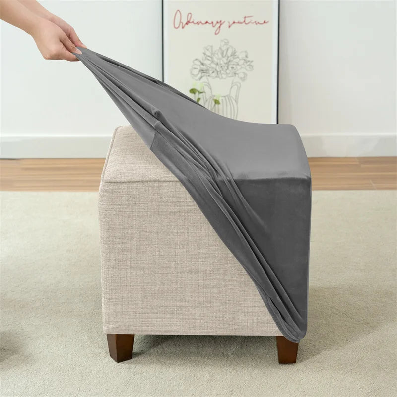 A gray velvet ottoman slipcover is being draped over a light-colored upholstered furniture piece.