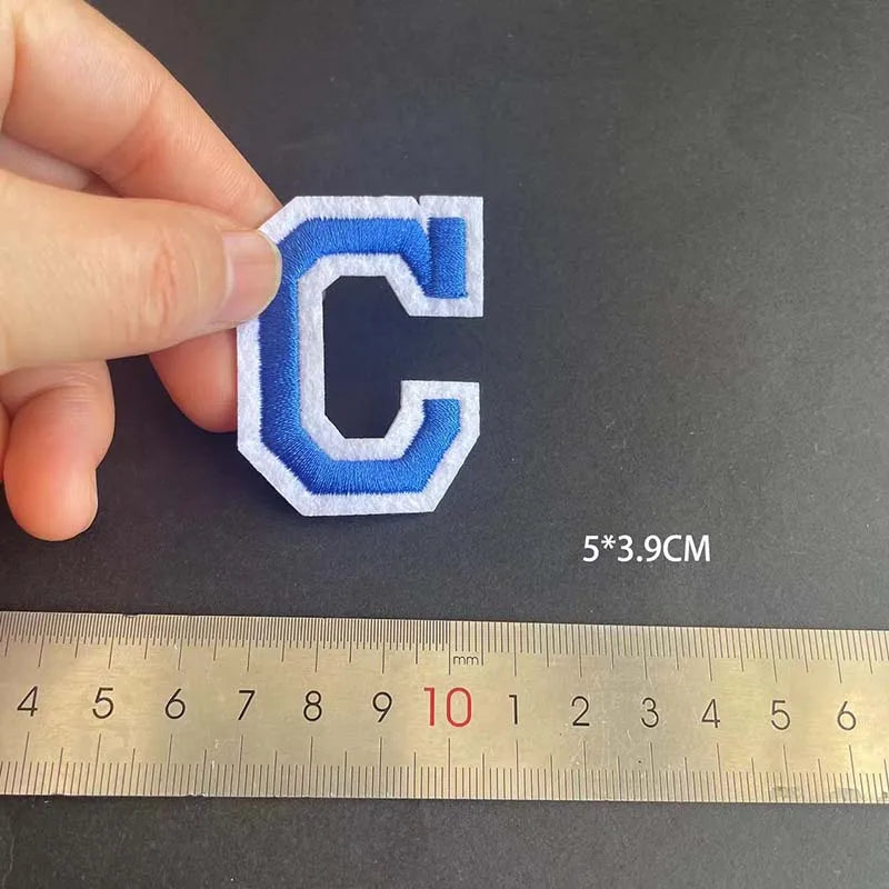 A hand holds a white and blue embroidered letter C patch, measuring 5x3.9 cm against a ruler.