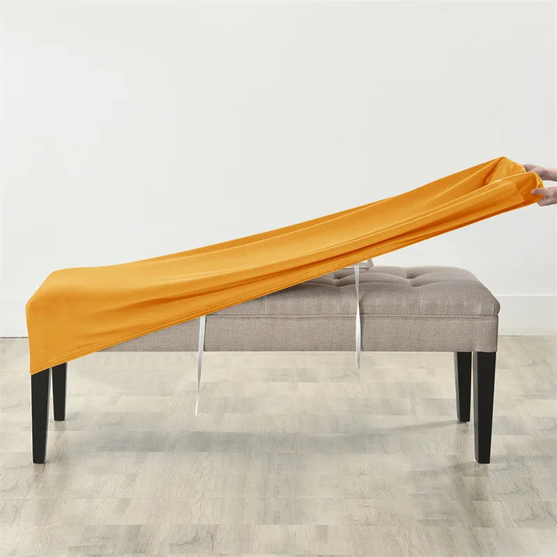 An orange stretchy velvet bench slipcover draped over a gray upholstered bench.