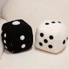 A pair of oversized dice plush pillows, one black with white dots and one white with black dots, resting on a light fabric su