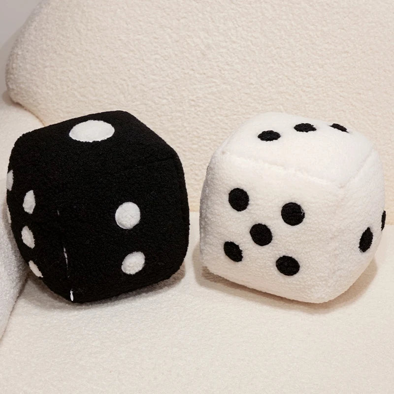 A pair of oversized dice plush pillows, one black with white dots and one white with black dots, resting on a light fabric su