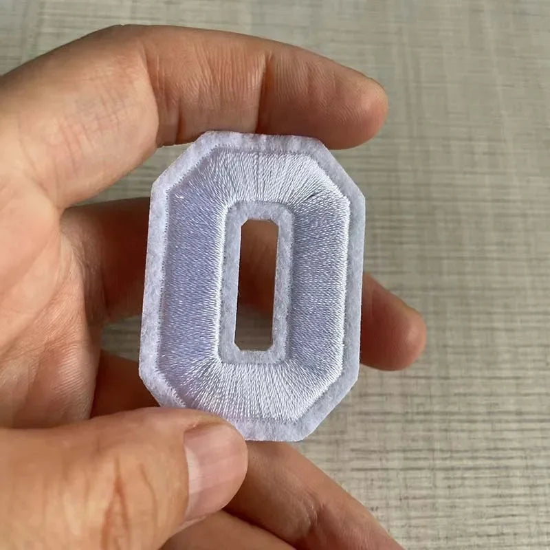 A hand holds a white, custom-shaped number patch with the numeral zero.