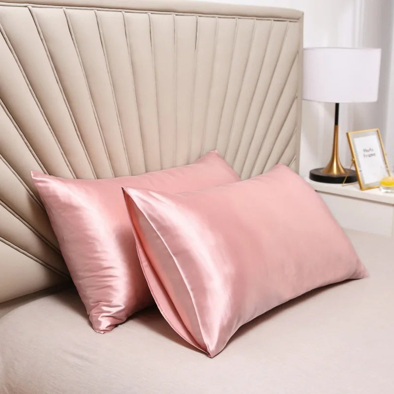 Pink satin silk pillow covers resting on a bed with a padded headboard and a bedside lamp.
