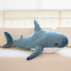 A soft blue giant shark plush toy lying on a light-colored sofa next to a window.