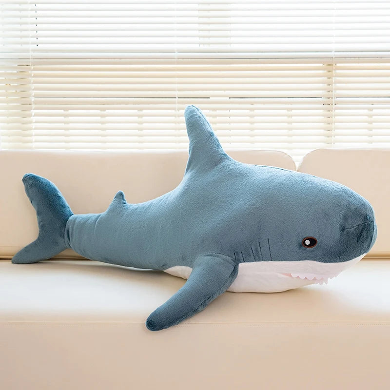 A soft blue giant shark plush toy lying on a light-colored sofa next to a window.