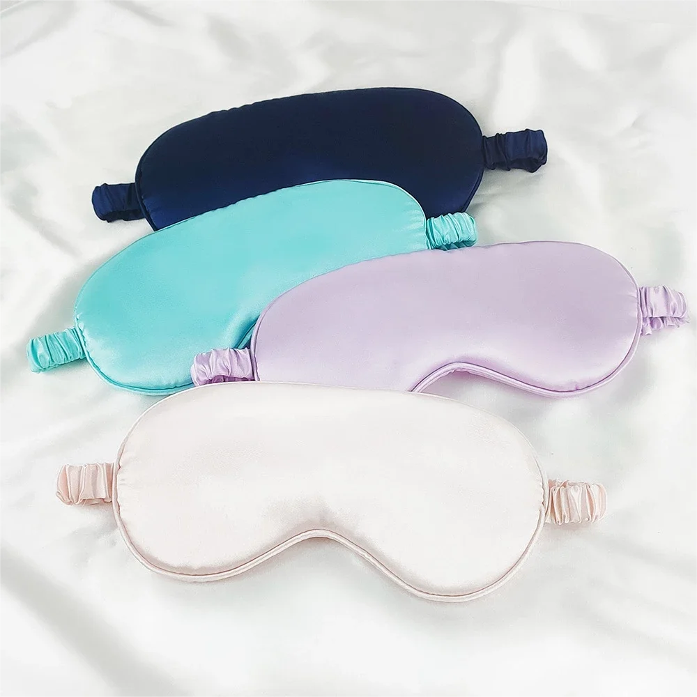 Three colored silk sleep masks in navy blue, turquoise, and lavender are displayed on a white surface.