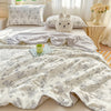 A light beige quilt blanket with a delicate gray floral pattern is draped over a bed, next to pillows and a small green cup.