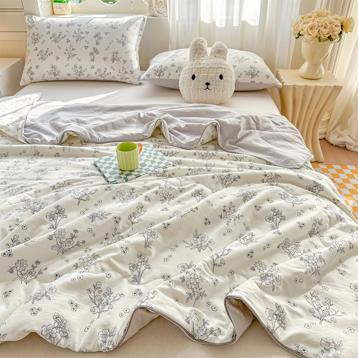 A light beige quilt blanket with a delicate gray floral pattern is draped over a bed, next to pillows and a small green cup.