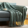 A teal knitted tassel throw blanket draped over a gray sofa with a white cup.