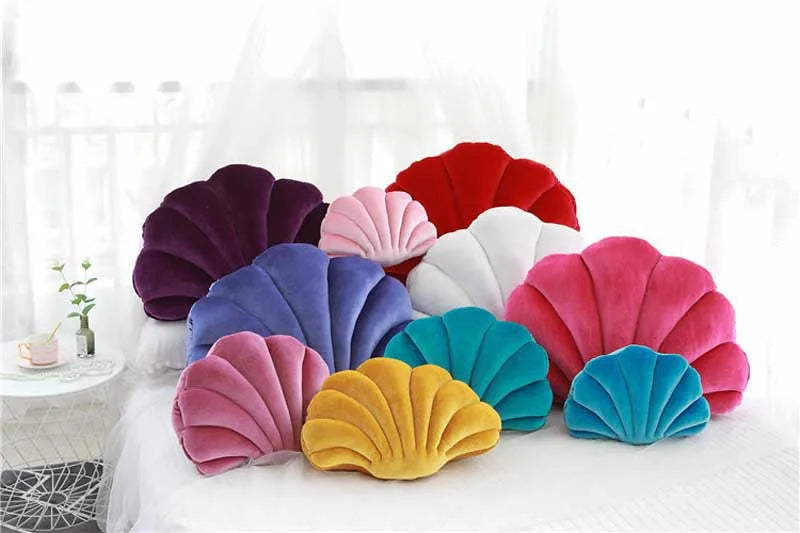 A collection of velvet stuffed sofa cushions shaped like decorative seashells in shades of purple, pink, red, white, and blue