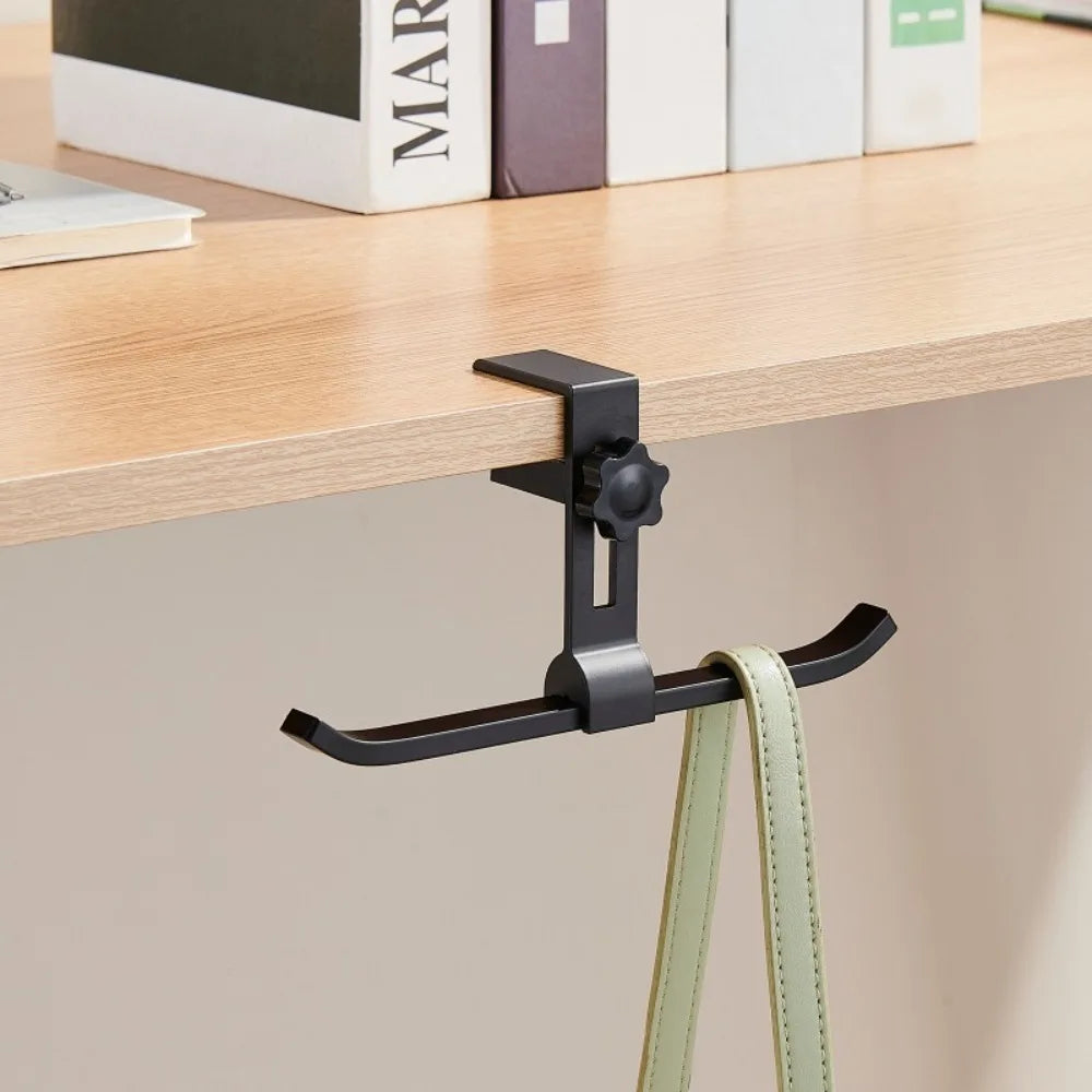 A black adjustable aluminum alloy desk hook with dual clips is hanging on a light wood shelf.