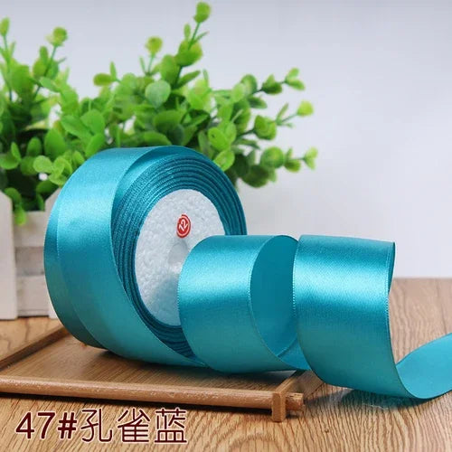 A roll of teal satin ribbon for gift wrapping and DIY crafts, resting on a wooden surface with green foliage in the backgroun