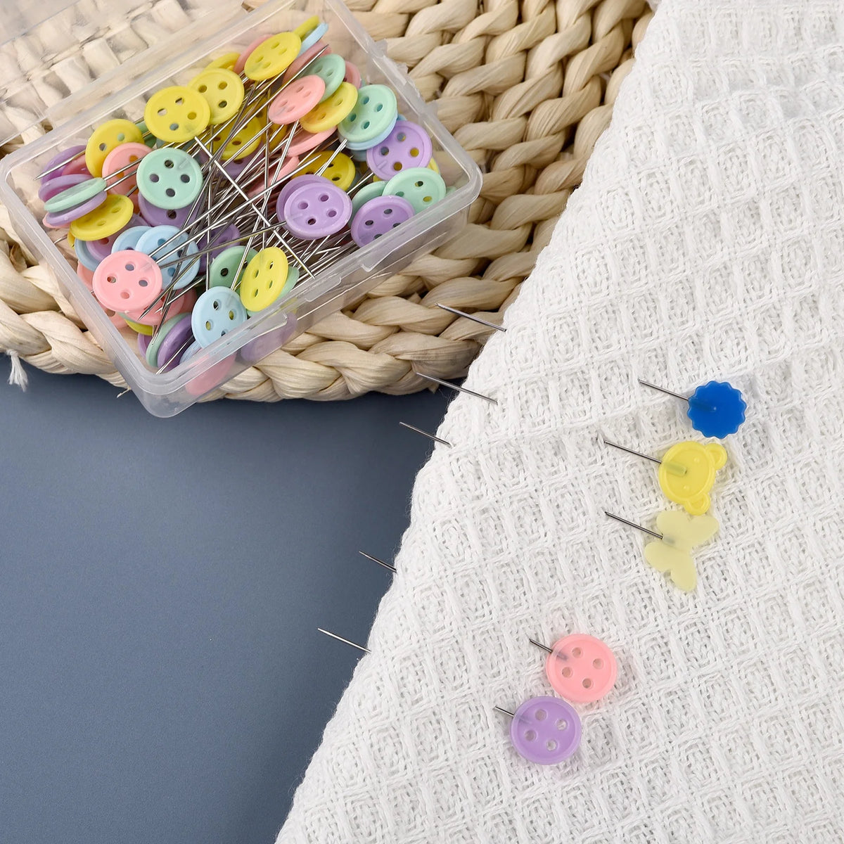 A clear plastic box filled with colorful nickel-plated dressmaking pins and needles resting on a woven mat.