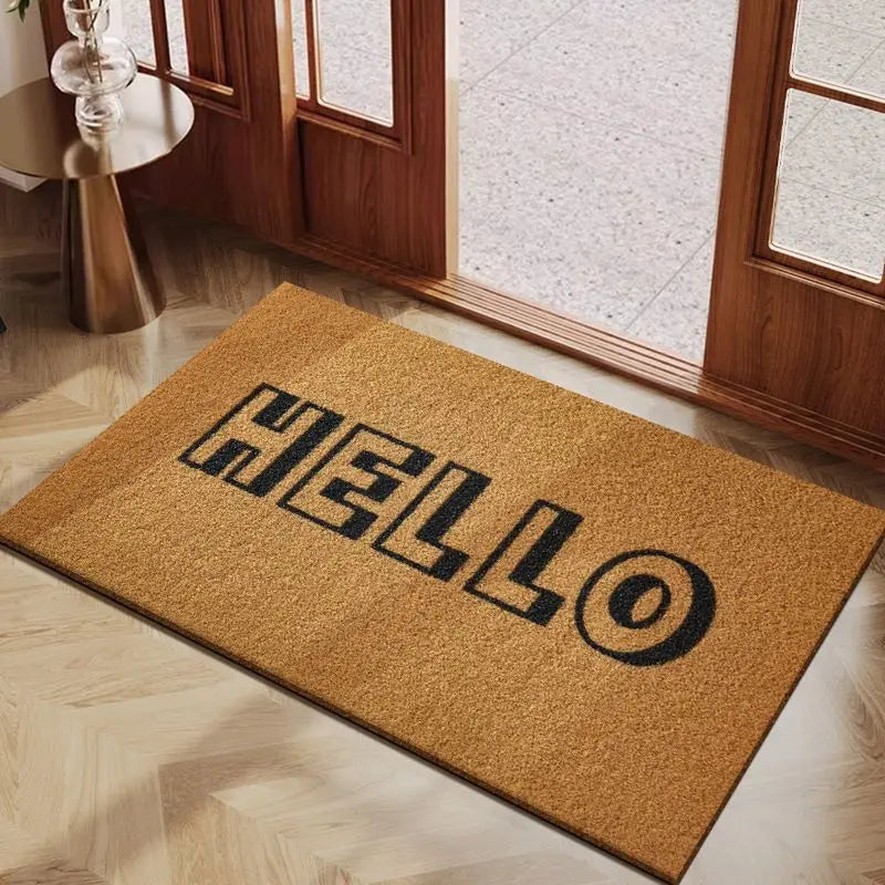A rectangular, golden-brown coconut fiber doormat with the word HELLO embossed on it, placed on a light wooden floor near a g