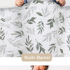 A baby is wrapped in a white muslin blanket featuring green leaf patterns.