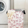A pink and white foldable canvas laundry basket decorated with a pattern of pink bows, placed next to a washing machine.