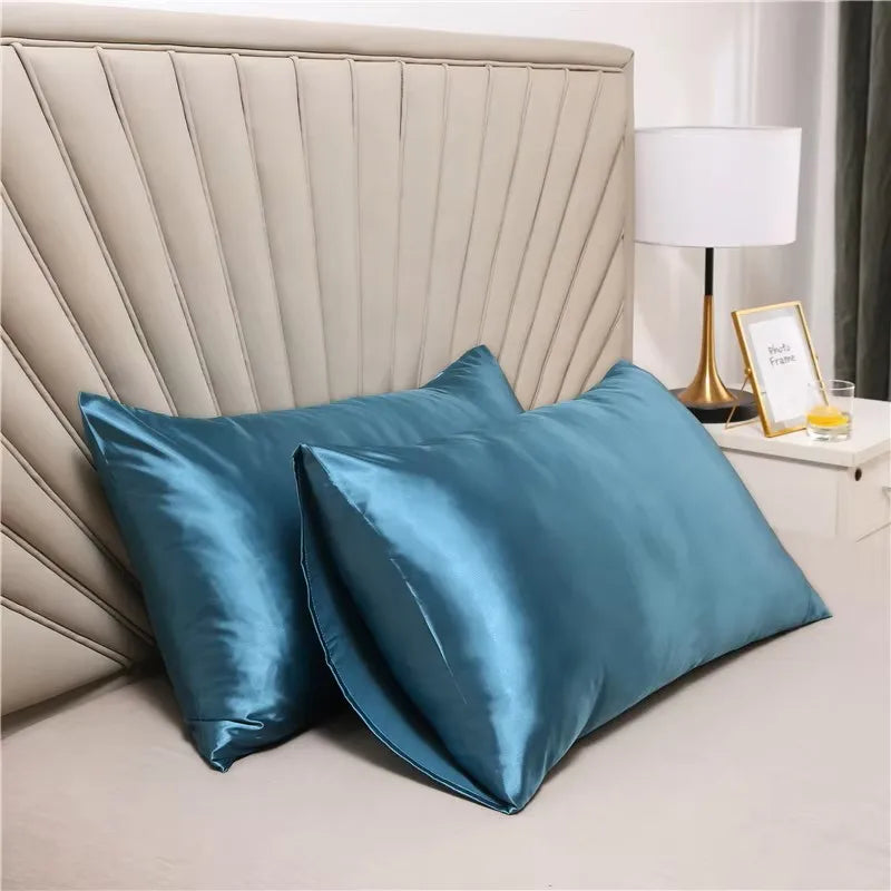 A teal silky satin pillowcase is displayed in front of a padded headboard and a lamp.