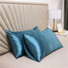 A teal silky satin pillowcase is displayed in front of a padded headboard and a lamp.