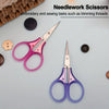 A set of needlework scissors in pink and purple colors with a protective cap shown on a light surface.