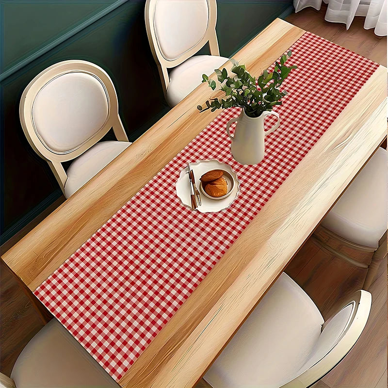 A light wood dining table set with a red and white checkered table runner, chairs, and a vase of greenery.
