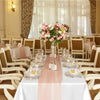 A white rectangular table set with a pink satin runner and glassware in an elegant dining room setting.