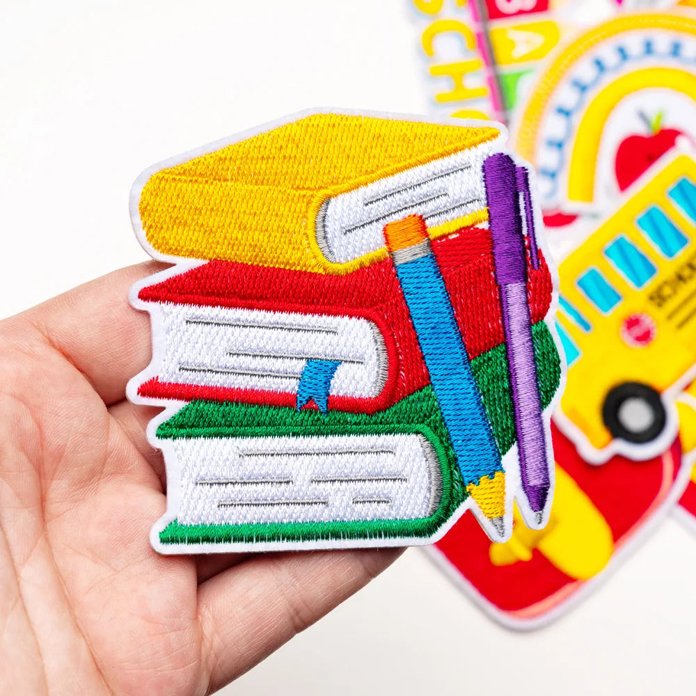 A hand holds a stack of colorful iron-on decorative patches featuring books, pencils, and a yellow school bus.