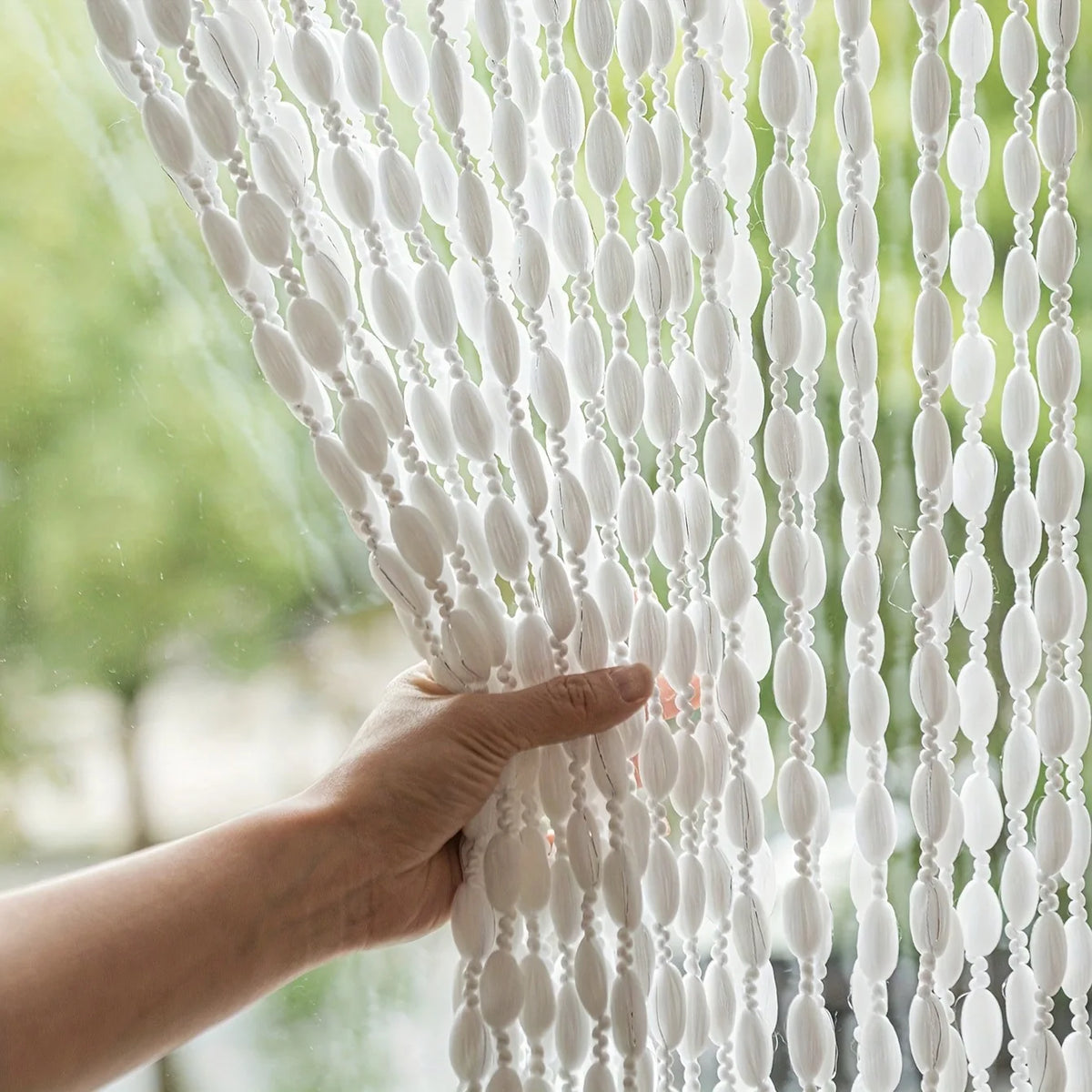 A hand touches a white decorative fabric door curtain with tassel trim, showing the sheer texture and vertical pleats.