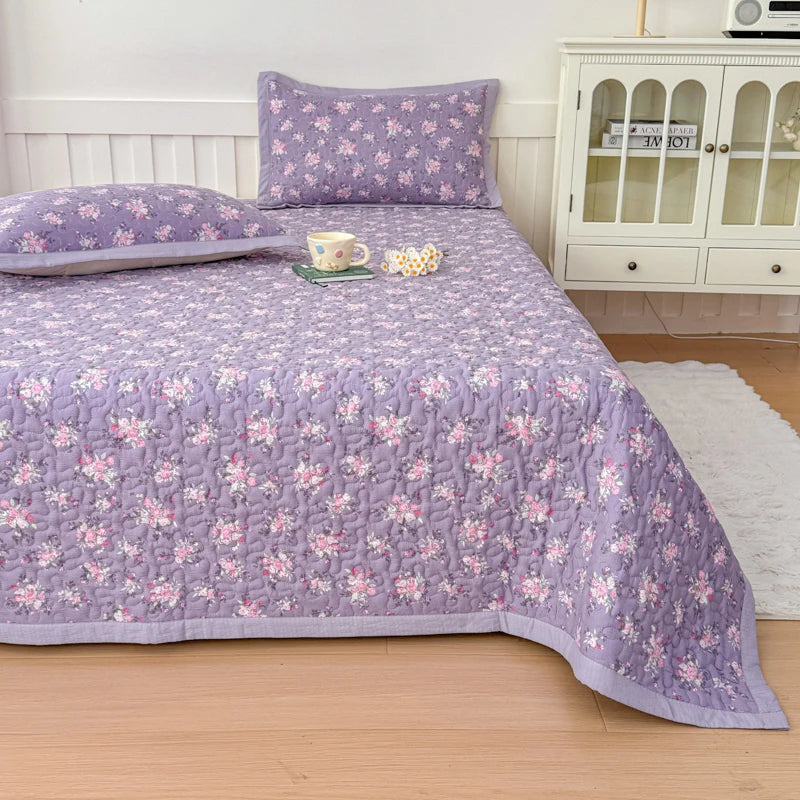 A purple floral quilted bedspread with matching pillows and a white cabinet in a bedroom setting.