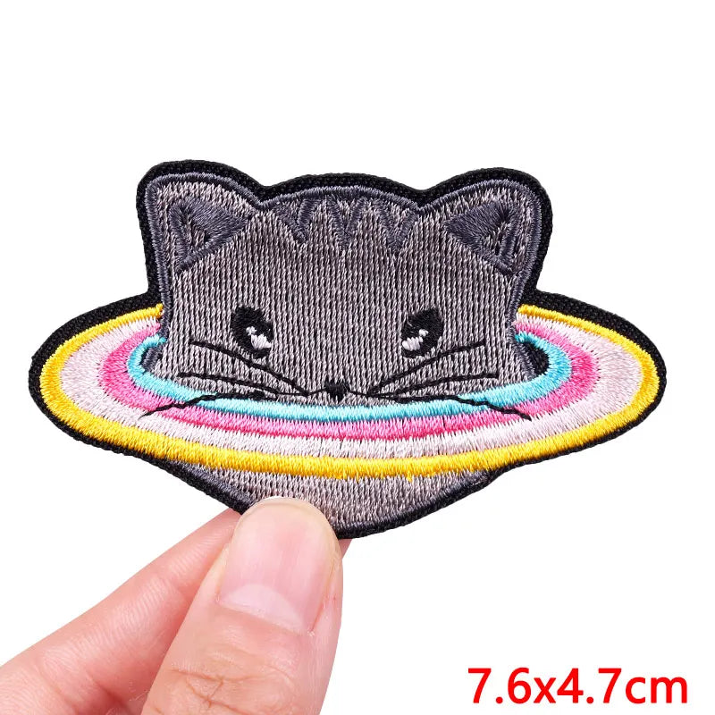 A gray embroidered cat patch with a colorful rainbow border being held by a hand.