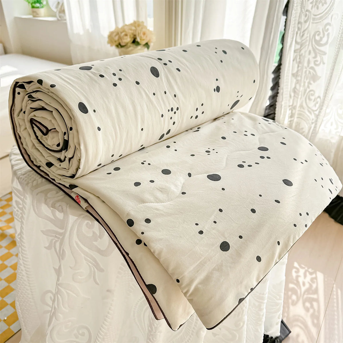 A cream-colored quilt blanket with black polka dots is rolled and draped on a white surface near sheer curtains.