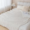 A white knitted blanket is draped over a bed, next to pillows and a shelf of rolled towels.